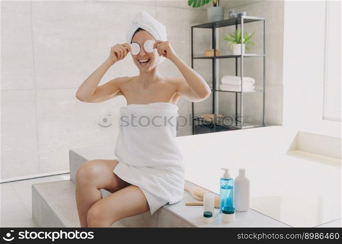 Happy young woman has fun applying face toner with cotton discs. Gorgeous european girl wrapped in towel after bathing. Lady is sitting on bathtub and relaxing. Woman takes shower at home.. Happy woman has fun applying face toner with cotton discs. Lady sitting on bathtub and relaxing.