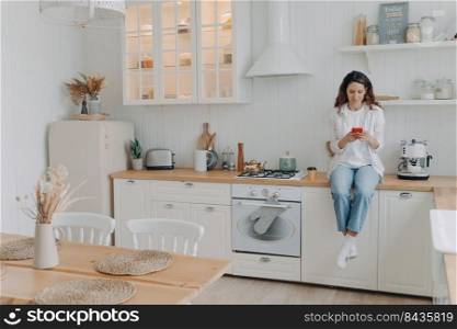 Happy young woman has message on phone. European housewife has leisure and sitting on worktop at kitchen at her apartment. Girl in casual outfit at home. Modern luxurious scandinavian interior.. Happy young woman has message on phone. Girl sitting on worktop at kitchen at her apartment