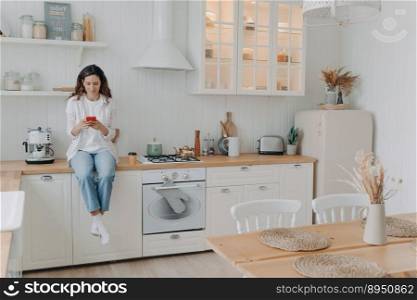 Happy young woman has message on phone. European housewife has leisure and sitting on worktop at kitchen at her apartment. Girl in casual outfit at home. Modern luxurious scandinavian interior.. Happy young woman has message on phone. Girl sitting on worktop at kitchen at her apartment