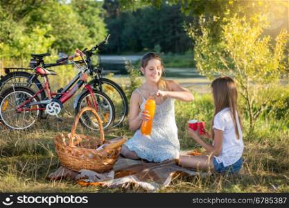Happy young woman having picnic by the river with 10 years old daughter. Two bicycles on the background