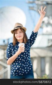 Happy young woman holding a takeaway coffee and showing three-finger gesture against background of parabolic satellite dishes that receive wireless signals from satellites.