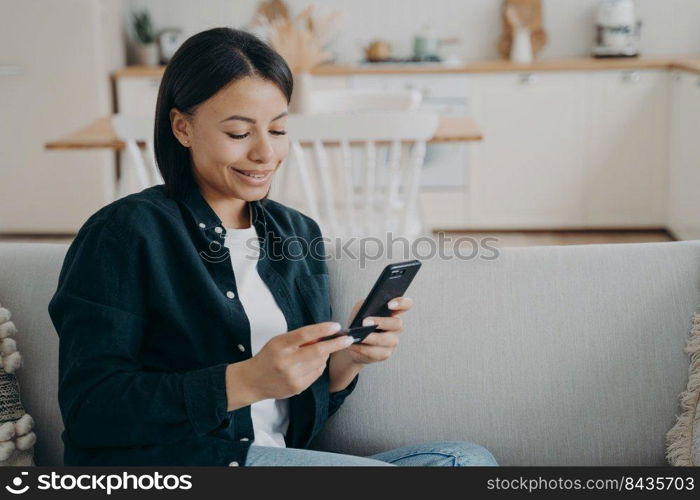 Happy young woman holding bank credit card, smartphone, entering information in online store app, purchasing, making successful cashless payment for internet services, sitting on sofa at home.. Woman holding bank credit card, smartphone makes successful cashless payment sitting on sofa at home