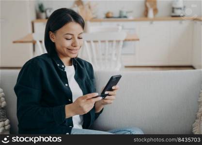 Happy young woman holding bank credit card, smartphone, entering information in online store app, purchasing, making successful cashless payment for internet services, sitting on sofa at home.. Woman holding bank credit card, smartphone makes successful cashless payment sitting on sofa at home