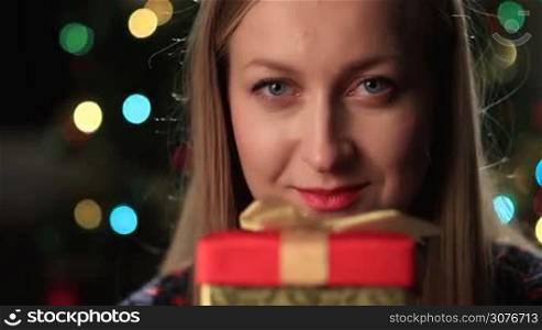 Happy young woman holding christmas gift box in front of face over colorful bokeh circles of xmas lights on tree background. Focus on woman&acute;s deep blue eyes.