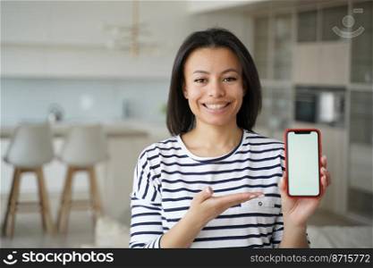Happy young woman holding phone with copy space, advertising new mobile apps. Smiling modern female showing smartphone with mockup empty screen, looking at camera, standing at home. Online store offer. New mobile apps. Smiling female showing phone with mockup empty screen at home. Online store offer