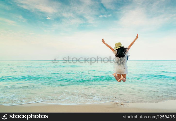 Happy young woman in casual style fashion and straw hat jumping at sand beach. Relaxing, fun, and enjoy holiday at tropical paradise beach with blue sky and white clouds. Girl in summer vacation.