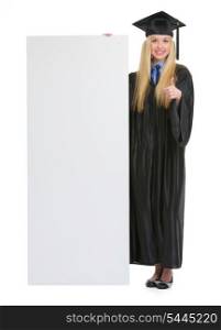 Happy young woman in graduation gown showing blank billboard and thumbs up