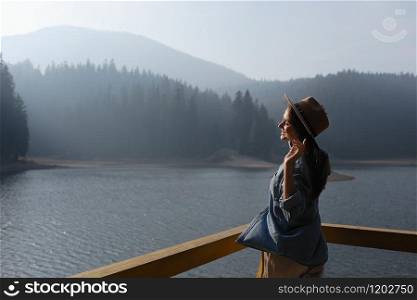 happy young woman in hat enjoys lake view in mountains . Relaxing moments in forest. Back view of stylish girl enjoys the freshness outdoor. Freedom, people, lifestyle, travel and vacation