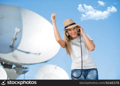 Happy young woman in hat listening to the music in vintage music headphones and dancing against background of satellite dish that receives wireless signals from satellites.