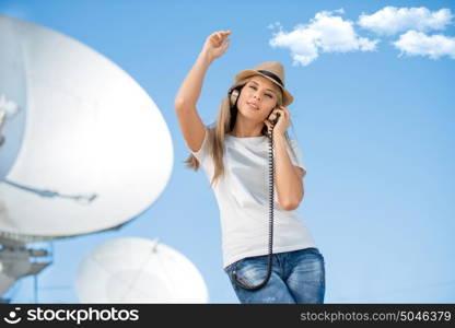 Happy young woman in hat listening to the music in vintage music headphones and dancing against background of satellite dish that receives wireless signals from satellites.