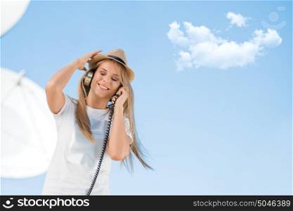 Happy young woman in hat listening to the music in vintage music headphones and dancing against background of satellite dish that receives wireless signals from satellites.
