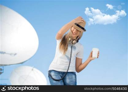 Happy young woman in hat wearing vintage music headphones around her neck, drinking takeaway coffee and posing against background of parabolic satellite dish that receives wireless signals from satellites.