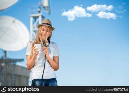 Happy young woman in hat wearing vintage music headphones around her neck, drinking takeaway coffee and posing against background of parabolic satellite dish that receives wireless signals from satellites.