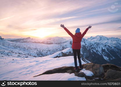 Happy young woman in snowy mountains at sunset in winter. Beautiful slim girl on the mountain peak with raised up arms, snow covered rocks and colorful sky with clouds. Travel in Dolomites. Tourism