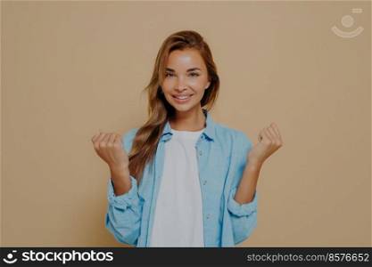 Happy young woman in stylish casual outfit holding hands in clenched fists, showing excitement and hope for best, smiling at camera while standing against beige background. People and success. Young woman holding hands in fists on beige background