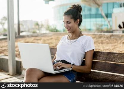 Happy young woman in summer outfit smiling and browsing social media on modern laptop while sitting on wooden bench on street. Smiling woman using laptop on bench