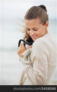 Happy young woman in sweater on beach with cup of hot beverage