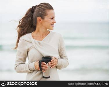 Happy young woman in sweater on beach with cup of hot beverage looking on copy space