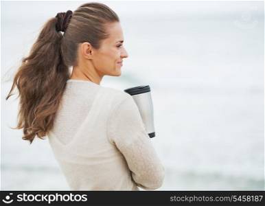 Happy young woman in sweater on beach with cup of hot beverage . rear view