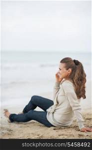 Happy young woman in sweater sitting on lonely beach and talking cell phone