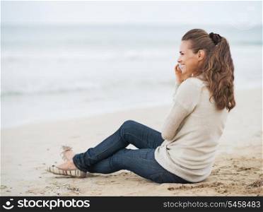 Happy young woman in sweater sitting on lonely beach and talking mobile phone