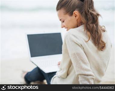 Happy young woman in sweater sitting on lonely beach and using laptop