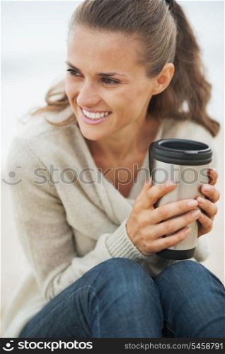 Happy young woman in sweater sitting on lonely beach with cup of hot beverage
