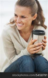 Happy young woman in sweater sitting on lonely beach with cup of hot beverage