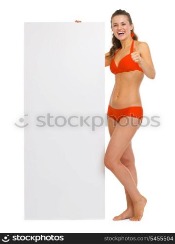 Happy young woman in swimsuit showing blank billboard and thumbs up