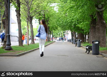 Happy young woman in white pants and a blue shirt walking in the summer park