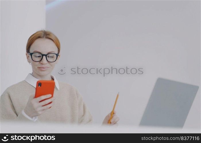 Happy young woman is chatting on phone while studying. Girl is sending messages while holding pencil. Browsing through internet, having fun and smiling. Being distracted at online study concept.. Happy young woman is chatting on phone having fun and smiling. Distracting at study concept.