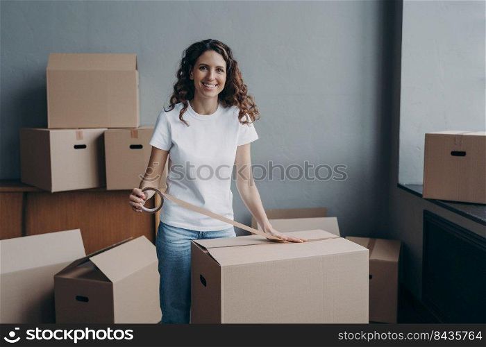 Happy young woman is packing boxes with duct tape. Attractive lady is wrapping cardboard boxes with packing tape. Moving service worker preparing cargo for shipping and storage.. Happy young woman is packing boxes with duct tape. Moving service worker preparing cargo.