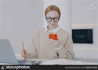 Happy young woman is texting at workplace in office. Girl is sending messages at her desk and playing with smartphone. Student is having fun. Being distracted at work concept.. Happy young woman is texting at workplace in office. Girl is sending messages at her desk.