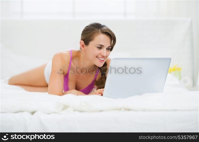 Happy young woman laying on bed and using laptop