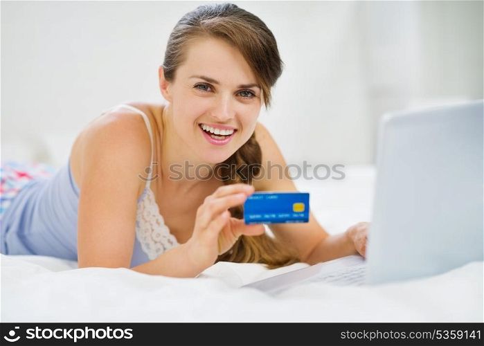Happy young woman laying on bed with credit card and laptop
