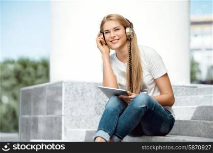 Happy young woman listening to the music in vintage music headphones, surfing internet on a tablet pc and sitting on stairs against urban city background.