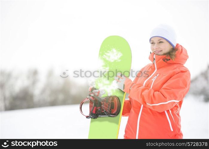 Happy young woman looking away while holding snowboard in snow