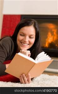 Happy young woman lying by fireplace on carpet reading book
