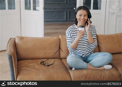 Happy young woman making phone call, talking, sitting on sofa with paper cup of coffee at home. Smiling female enjoying pleasant mobile conversation, holding takeaway drink in living room.. Happy girl makes phone call, sitting on sofa with coffee cup at home. Pleasant mobile conversation