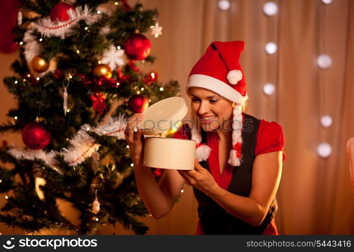 Happy young woman near Christmas tree looking inside present box&#xA;