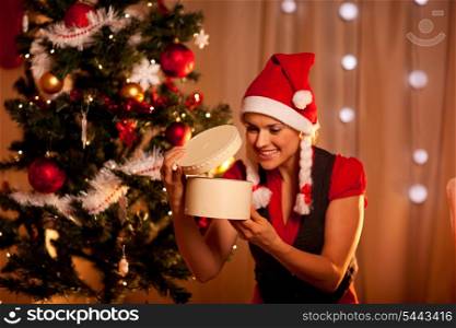 Happy young woman near Christmas tree looking inside present box&#xA;