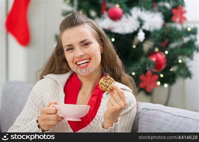 Happy young woman near Christmas tree with cup and cookie
