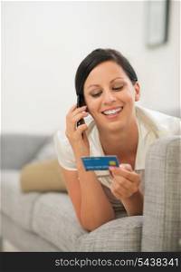 Happy young woman on sofa holding credit card and speaking mobile phone
