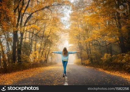 Happy young woman on the road in beautiful autumn forest in fog at sunset. Landscape with alone girl, road, orange trees. Empty roadway through foggy woodland in fall. Travel and adventure in Europe. Young woman on the road in autumn forest at sunset
