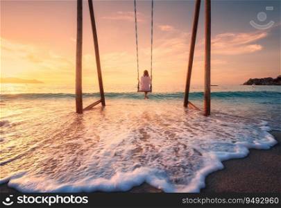 Happy young woman on wooden swing in water, beautiful blue sea with waves, sandy beach, orange sky at sunset. Summer holiday in Oludeniz, Turkey. Girl ride on a swing on sea coast, clear water. Travel
