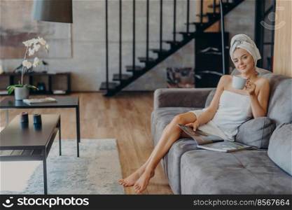 Happy young woman poses wrapped in bath towel, drinks aromatic coffee and reads magazine, sits on comfortable sofa, poses in living room at home. Domestic atmoshpere, leisure time, lifestyle