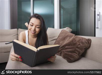 Happy young woman reading book while lying on sofa