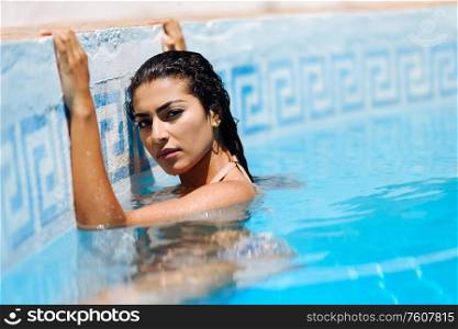 Happy young woman relaxing in swimming pool. Beautiful girl with healthy tanned skin and wet hair enjoying Summer Sun at pool edge.. Beautiful Arab woman relaxing in swimming pool.