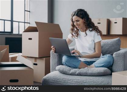 Happy young woman selling or purchasing apartment online. Spanish girl among cardboard boxes is using computer and smiling. Investment and internet banking.. Happy young woman selling or purchasing apartment online. Investment and internet banking.