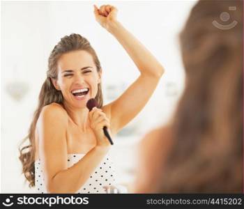 Happy young woman singing in makeup brush in bathroom